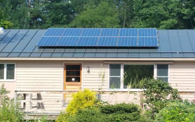 Solar panels on metal roof of one story house with deck and bushes in front